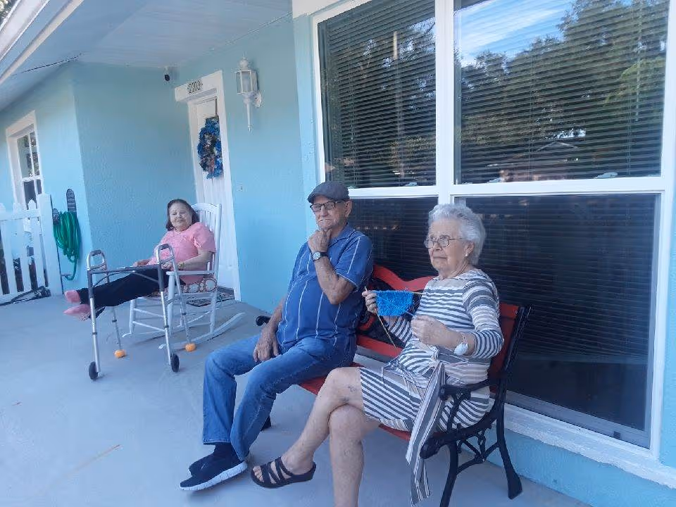 Three elderly individuals sitting outside a light blue building. One woman in a pink shirt is seated on a rocking chair with a walker in front of her. A man in a blue striped shirt and cap is sitting on a red bench next to an elderly woman in a striped dress who is holding a blue face mask. The building has large windows and a white door with a wreath.