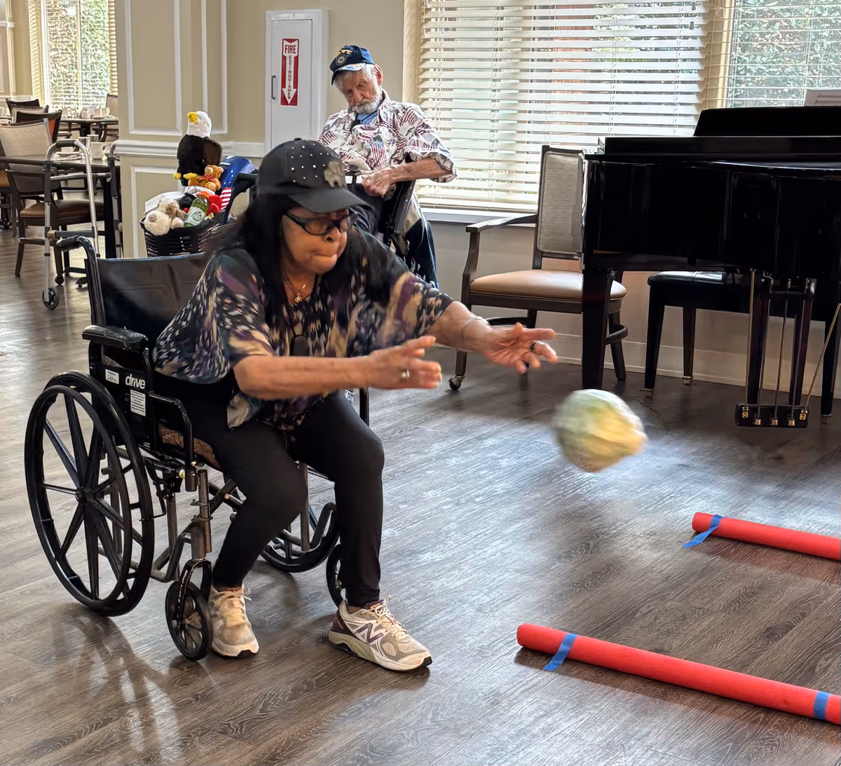A woman in a wheelchair reaches to toss a ball during a group activity in a senior living facility common room.