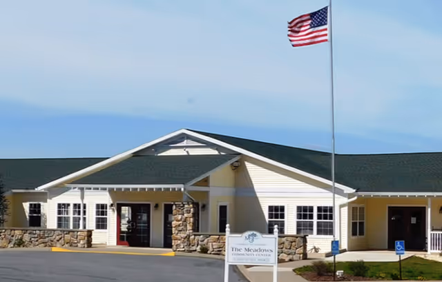 Exterior view of a single-story community center building with beige siding and stone accents. The building has a green roof and a covered entrance. An American flag flies on a tall flagpole in front of the building. There is a sign in front that reads 'The Meadows Community Center'.