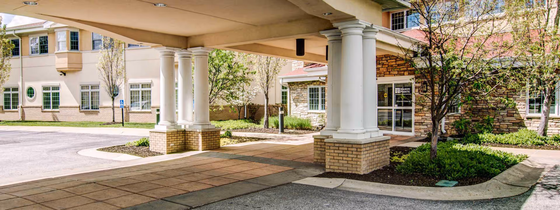 Covered porte-cochere with white columns and a driveway at the front entrance of the EverTrue Mason Pointe building, with landscaping and glass doors.