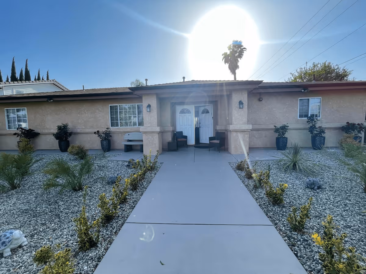 Front exterior view of a single-story building with a beige stucco finish, two white doors at the entrance, two chairs and a bench on the porch, surrounded by landscaped gravel and small plants under a clear blue sky with the sun shining brightly.