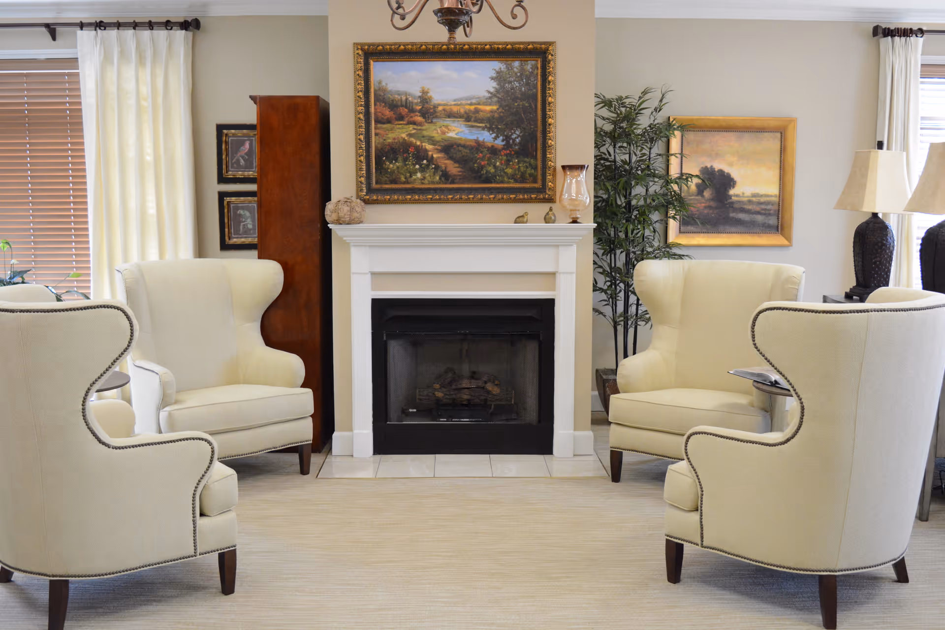 A cozy living room area with four cream-colored wingback chairs arranged around a white fireplace. Above the fireplace is a framed landscape painting. There are two additional framed artworks on the walls, a tall wooden cabinet, a potted plant, and two table lamps on a side table near a window with white curtains and wooden blinds.