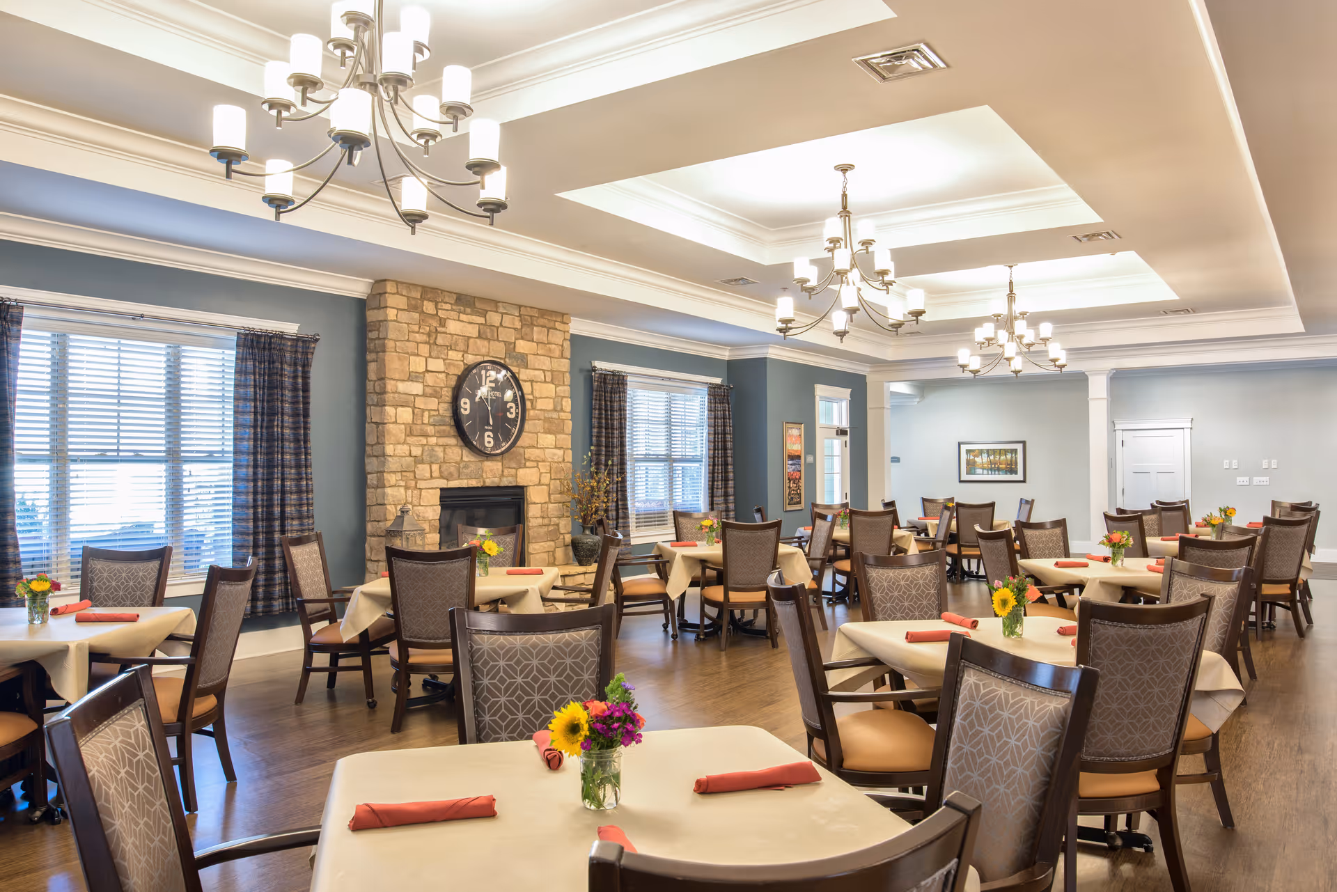 Bright, well-appointed dining room with tables set with napkins and flower centerpieces, chandeliers, and a stone fireplace.