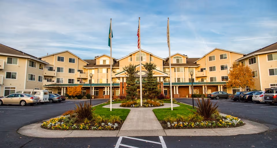 Front entrance of a multi-story senior living building with three flagpoles, landscaped island, and parking lot.