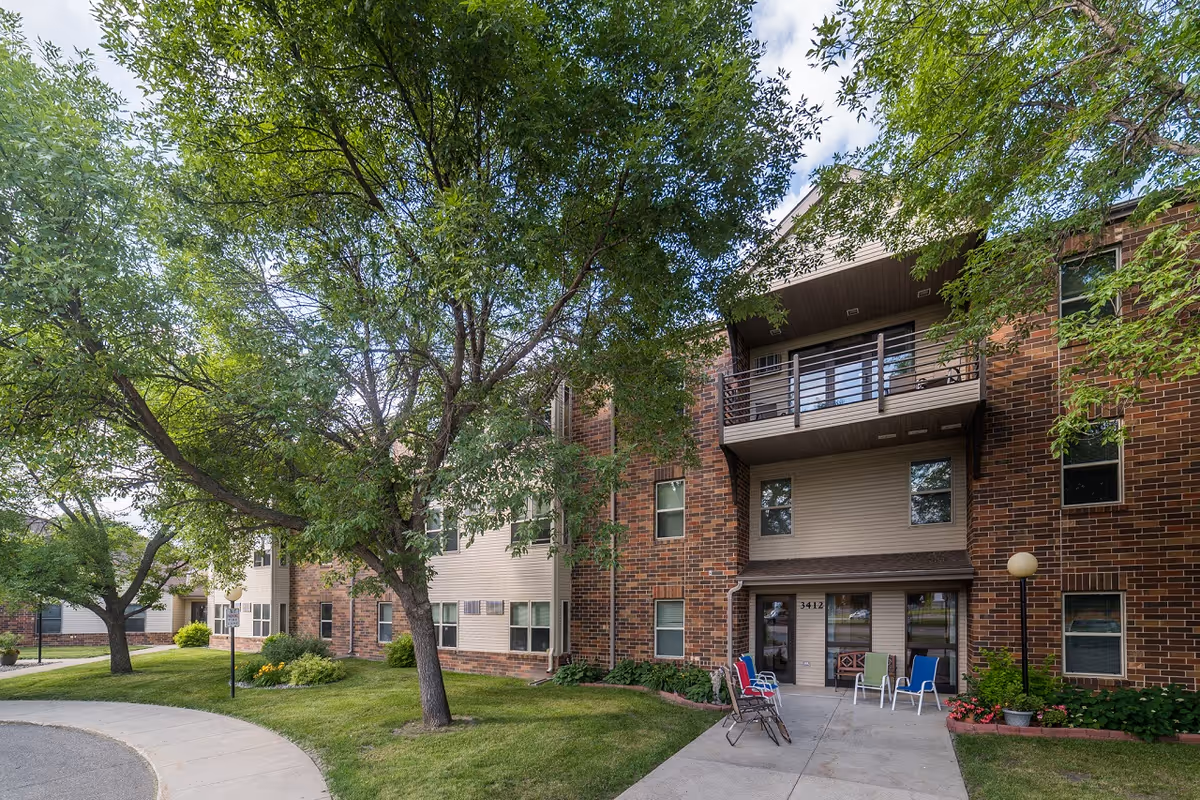 Exterior view of a multi-story senior living facility building named Carriage House with brick and beige siding, surrounded by green trees and a well-maintained lawn. Several colorful chairs are placed near the entrance under a covered balcony.