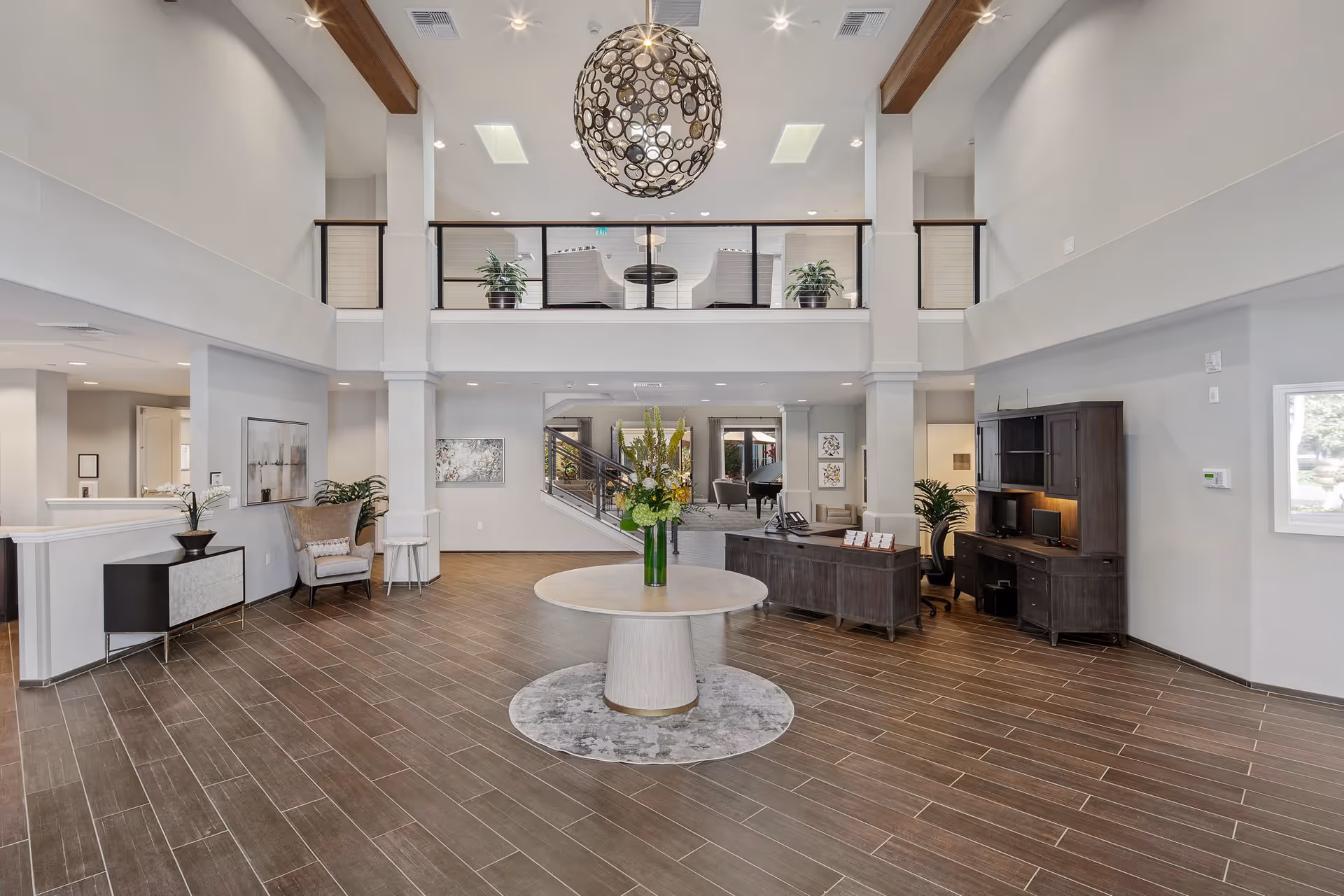 Spacious and modern lobby area with high ceilings, a round table with a flower arrangement in the center, a reception desk with a computer, comfortable seating areas, decorative plants, and a staircase leading to an upper level with additional seating.