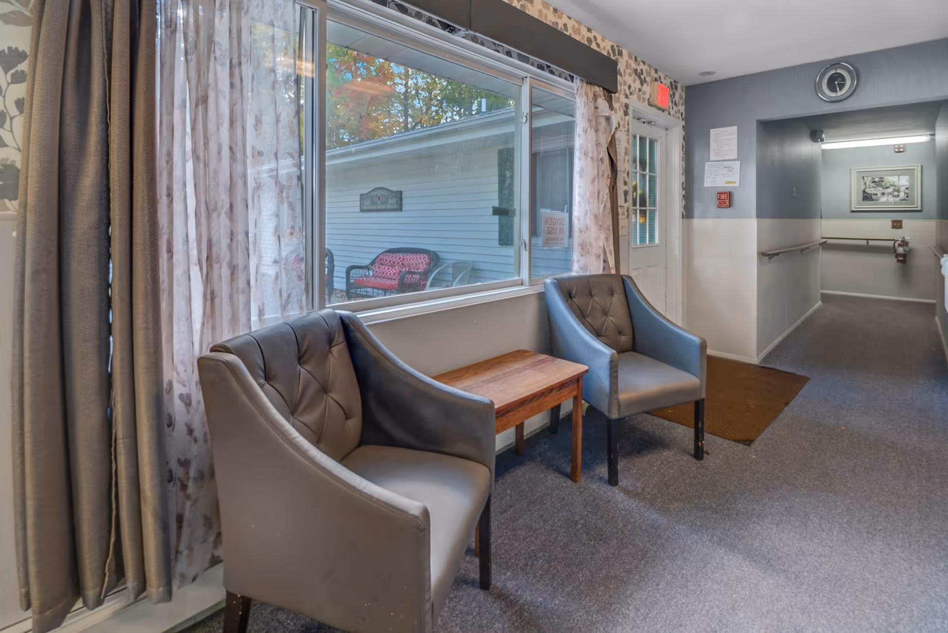 Small seating area in a facility hallway with two gray armchairs and a wooden side table beside a large window.