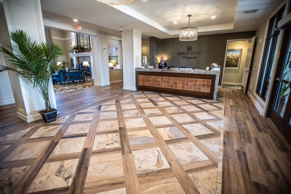 Reception area of Grand Living At Bridgewater featuring a front desk with a receptionist, decorative flooring with a wood and stone pattern, a large potted plant, and a seating area with blue chairs and a fireplace in the background.
