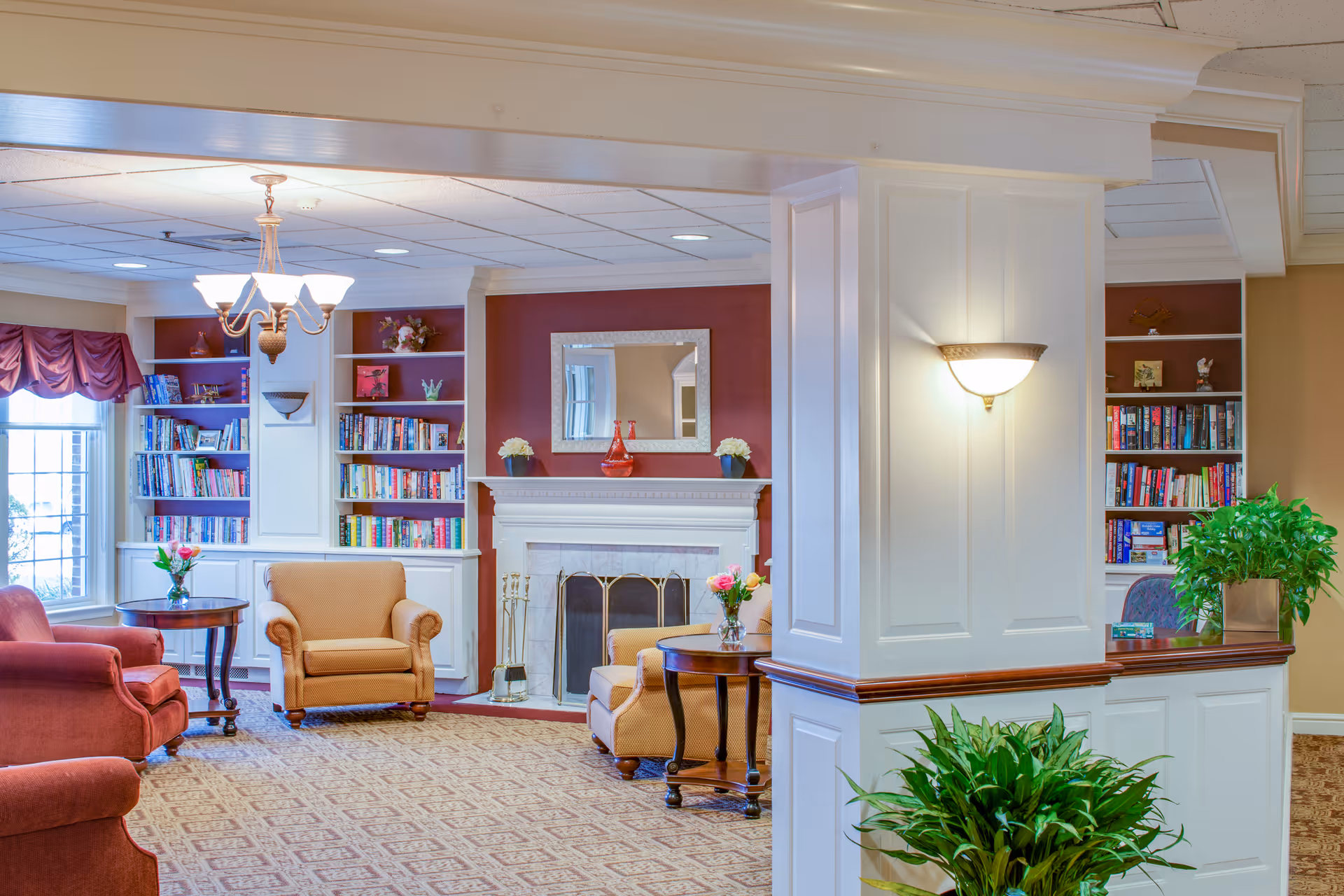 A cozy living room area in a senior living facility with comfortable armchairs, a fireplace with a mirror above it, built-in bookshelves filled with books, a chandelier, and decorative plants. The room has warm lighting and a patterned carpet.