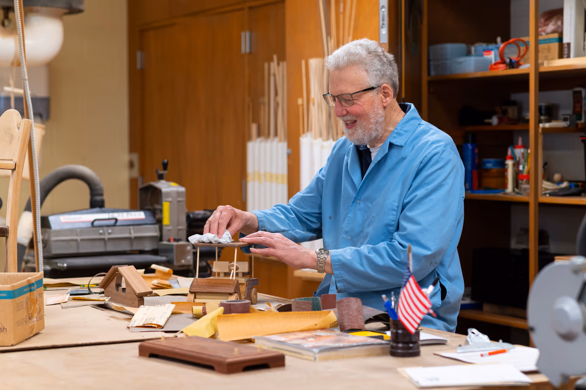 An elderly man wearing glasses and a blue work coat is working on a small wooden model in a workshop filled with woodworking tools and materials. He is smiling and appears focused on his craft.