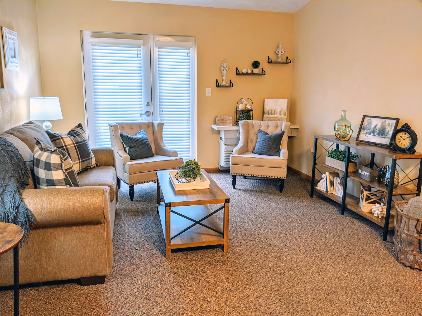 A cozy living room with beige walls and carpet. The room features a beige sofa with plaid and solid pillows, two beige armchairs with dark pillows, a wooden coffee table with a decorative plant, and a wooden shelving unit with various decorative items including a clock, framed pictures, and plants. There is a lamp on a small side table next to the sofa, and French doors with blinds letting in natural light.
