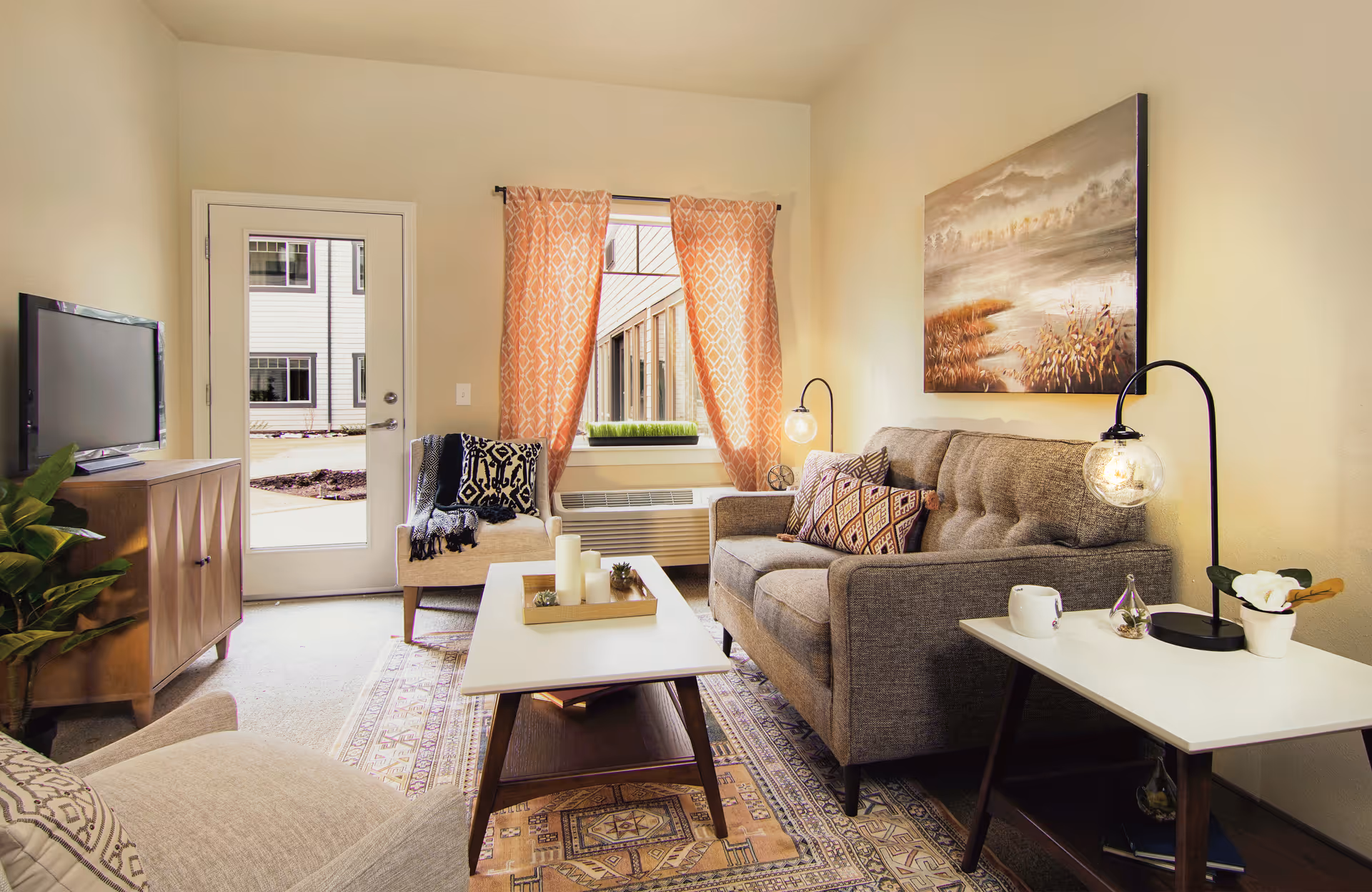 A cozy living room in a senior living community featuring a gray sofa with patterned cushions, a beige armchair with a black and white throw, a wooden coffee table with candles, a side table with a lamp and a small plant, a TV on a wooden stand, and a window with orange patterned curtains overlooking an outdoor courtyard.