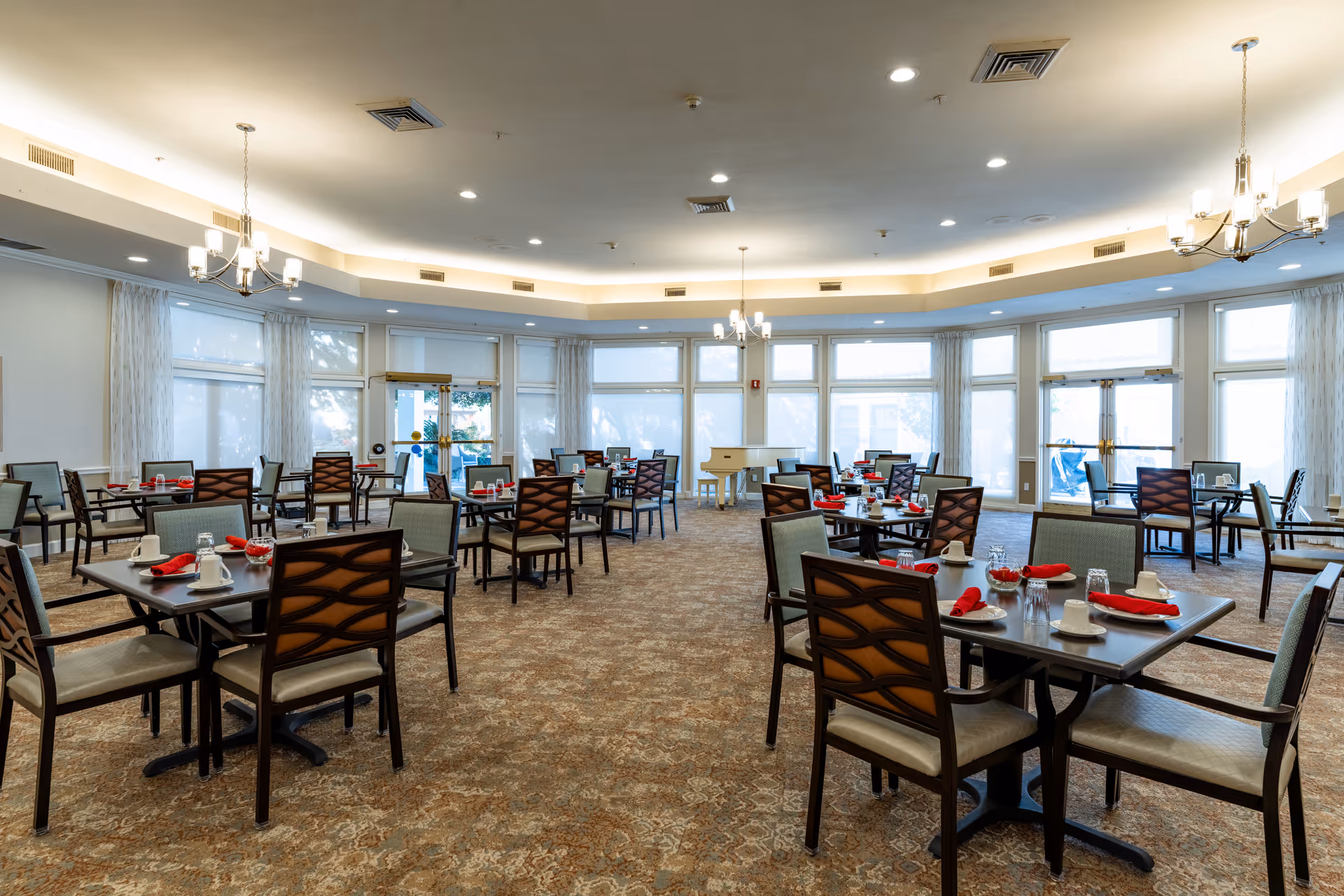 A spacious dining room with multiple tables set with cups, glasses, plates, and red napkins. The room has large windows with sheer curtains allowing natural light to fill the space. There are chandeliers hanging from the ceiling and a patterned carpet covering the floor.