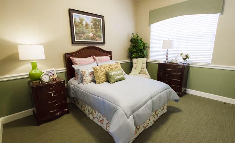 A cozy bedroom with a neatly made bed featuring multiple pillows, including one with the text 'welcome home'. The room has green and beige walls, a wooden headboard, two wooden nightstands with lamps, a framed landscape painting above the bed, a cushioned chair, a potted plant, and a window with blinds and a green valance.