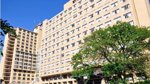 Exterior view of a large multi-story beige building with many windows under a clear blue sky. A green leafy tree is visible in the foreground.