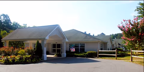 Exterior view of Cambridge Hills Assisted Living facility showing a single-story building with a covered entrance, surrounded by landscaped bushes, trees, and a wooden fence under a clear sky.