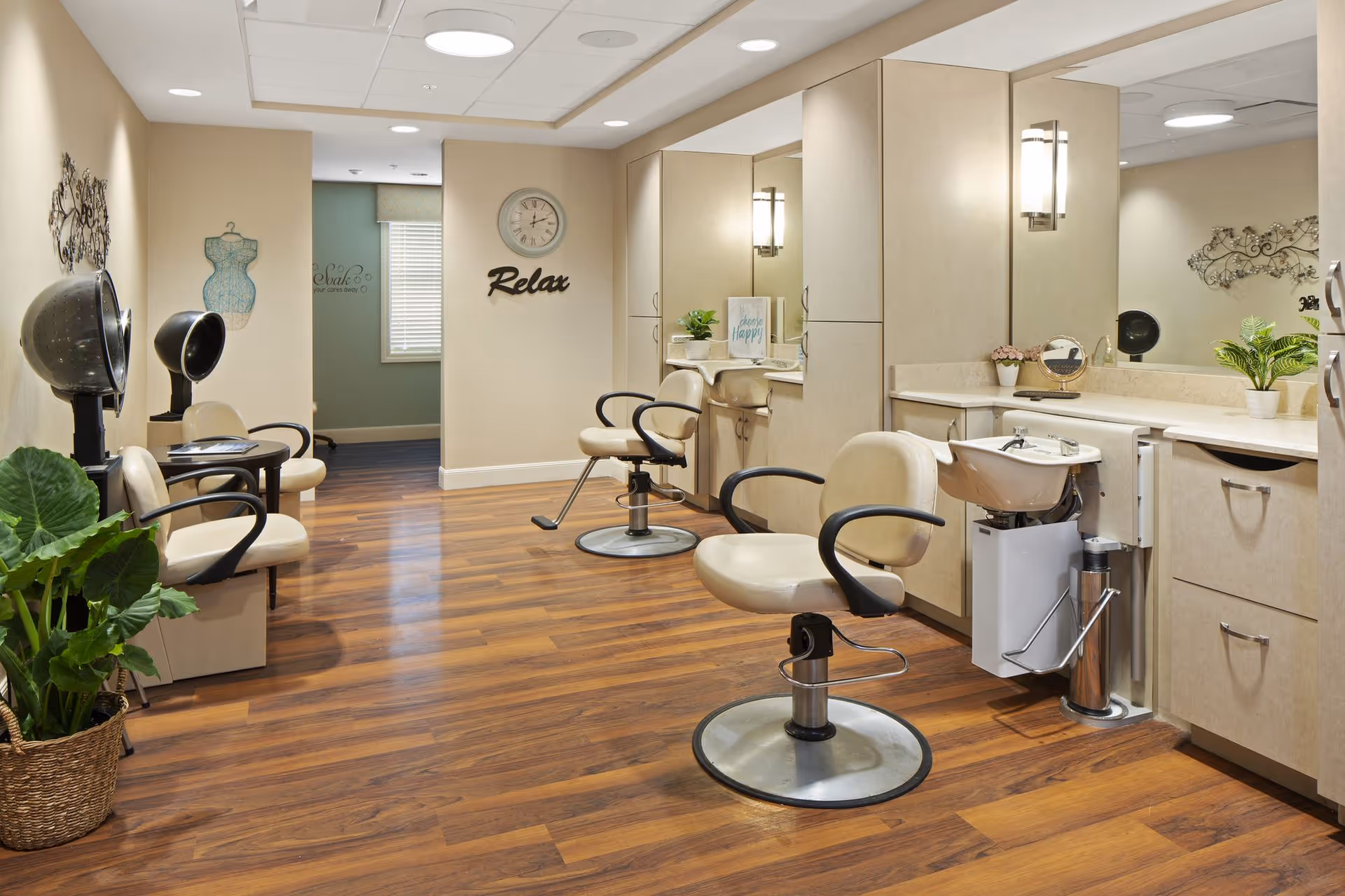 Interior of a salon area in a senior living facility with beige salon chairs, hair drying stations, a hair washing sink, wooden flooring, and decorative plants. The walls are light-colored with a clock and the word 'Relax' displayed.