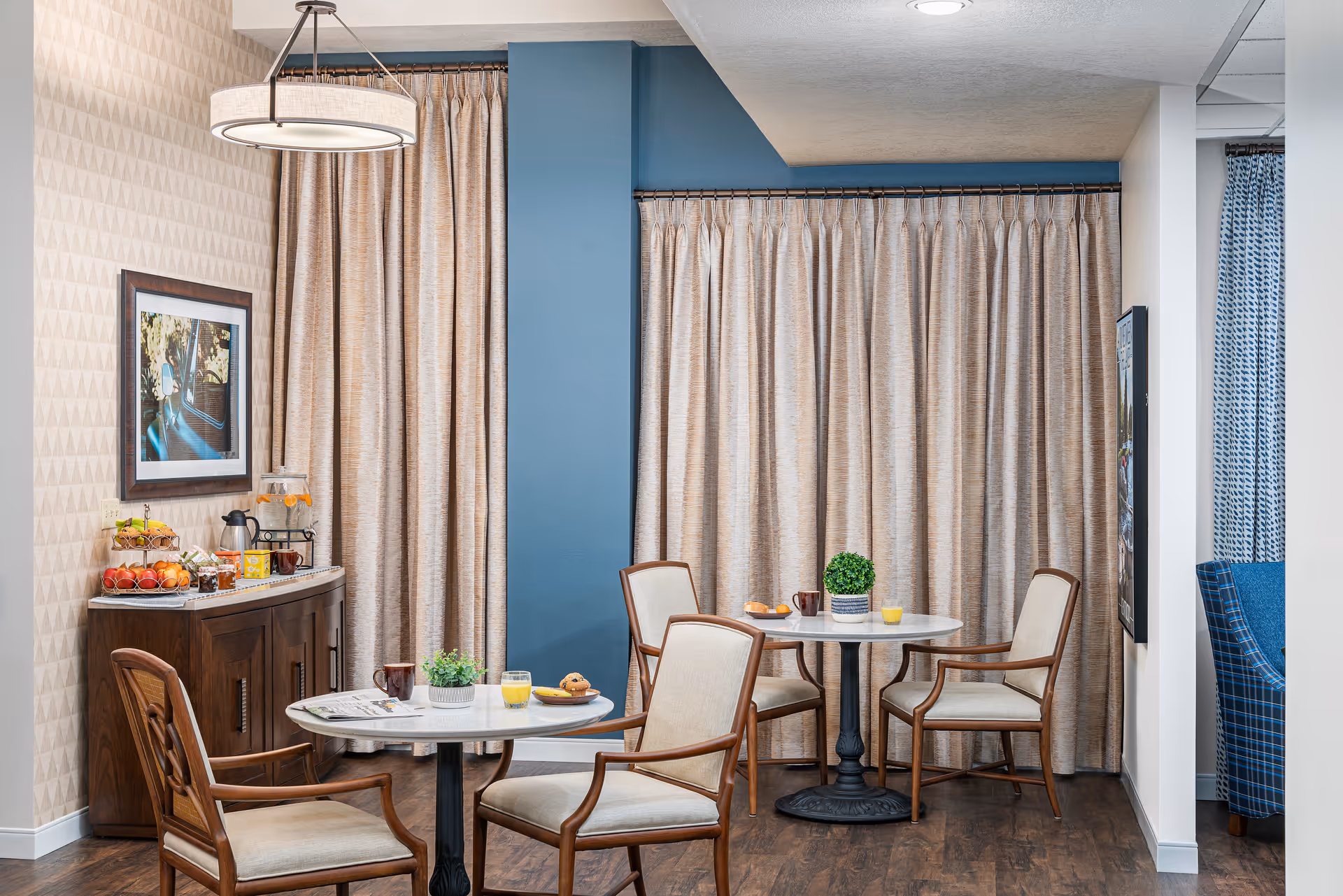 A cozy dining area with two round tables, each surrounded by four wooden chairs with beige cushions. On the tables are small plants, cups, and plates with pastries. Behind the tables are beige curtains covering the windows, and to the left is a wooden sideboard with a beverage dispenser, cups, and a fruit tray. The walls have a patterned wallpaper and a framed picture, and the floor is dark wood.