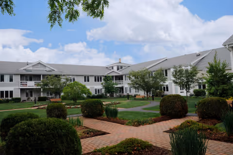 Sunlit courtyard with brick walkways, trimmed shrubs and lawns in front of a two-story white senior living building under a partly cloudy sky.