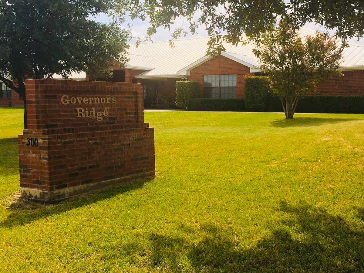 Brick sign reading 'Governors Ridge' on a green lawn with the retirement facility building visible behind it.
