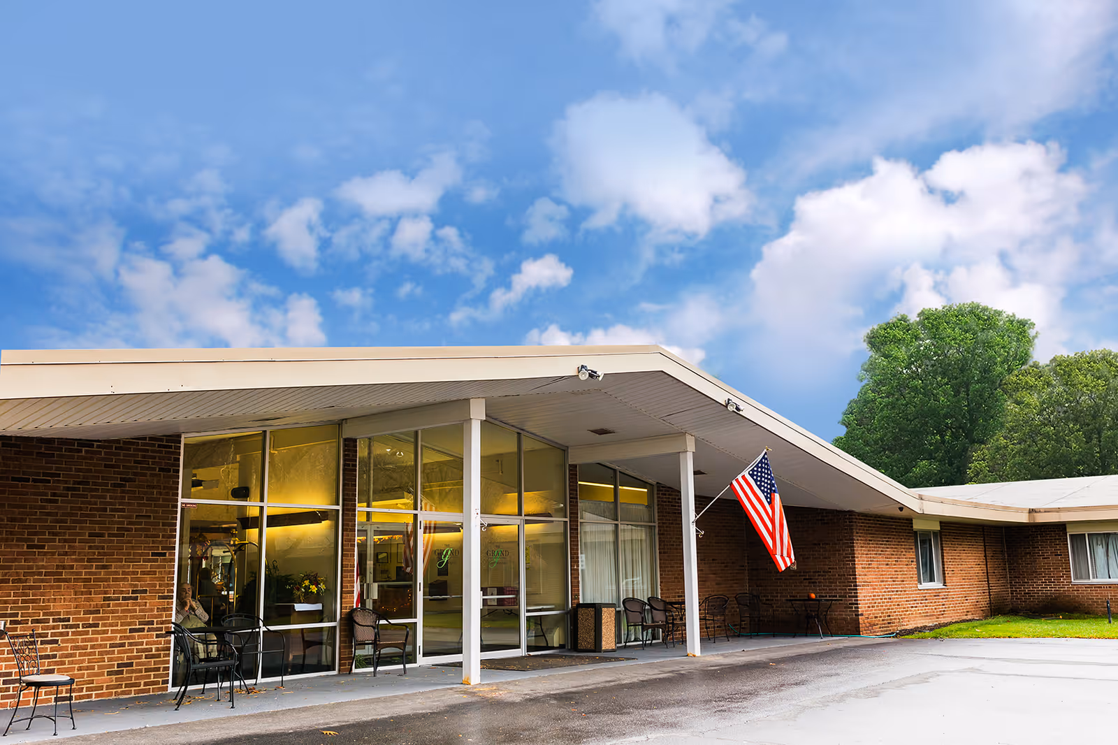 Exterior view of The Grand Rehabilitation and Nursing at Chittenango building with a covered entrance, large glass windows, several chairs outside, and an American flag mounted on a pole near the entrance. The sky is partly cloudy and there are green trees in the background.