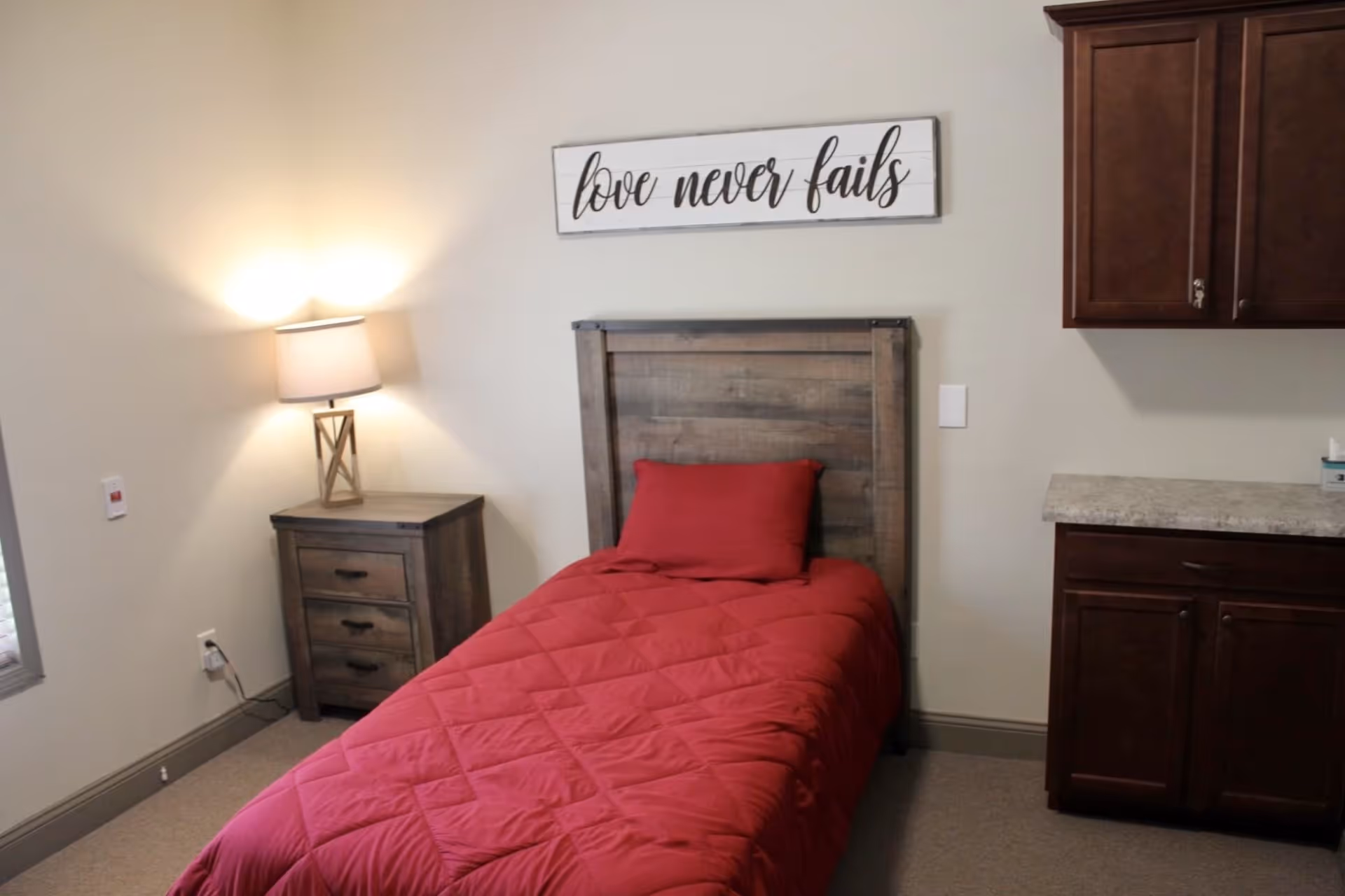 Small bedroom with a single bed dressed in a red comforter, a wooden headboard, nightstand with a lamp, cabinets, and a wall sign reading "love never fails".