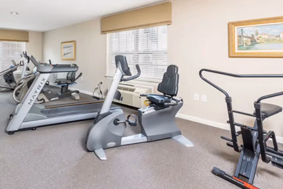 A fitness room with exercise equipment including a treadmill, a recumbent stationary bike, and a rowing machine. The room has beige walls, carpeted floor, two windows with blinds, and framed artwork on the walls.