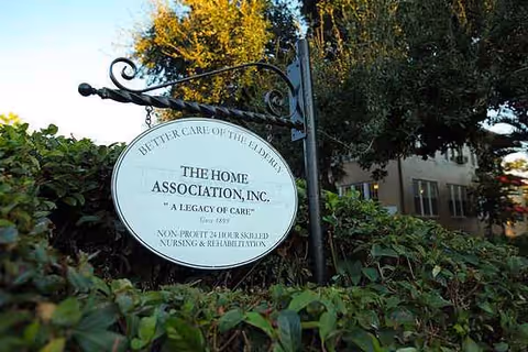 An outdoor sign for The Home Association, Inc., mounted near greenery by a building.