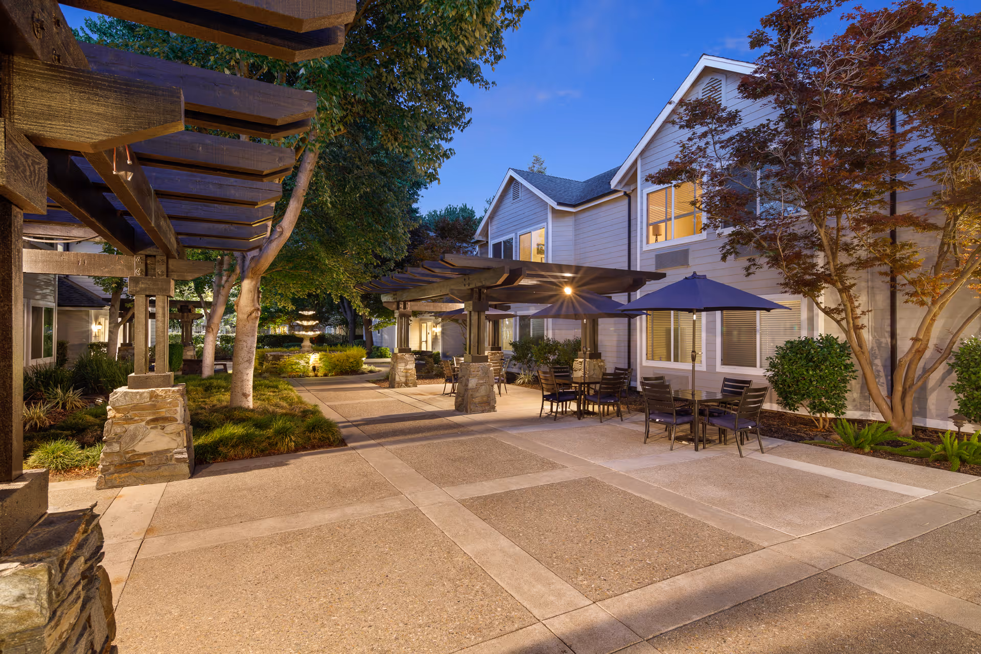 Outdoor patio area at dusk with tables, chairs, and umbrellas under wooden pergolas, surrounded by trees and shrubs, adjacent to a two-story building with lit windows.