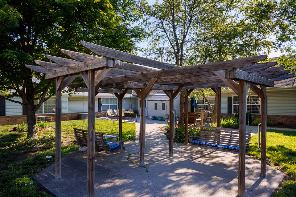 Outdoor seating area with a wooden pergola structure featuring two wooden swing benches with blue cushions. The area is surrounded by green grass, trees, and a building with windows and a brick foundation in the background.