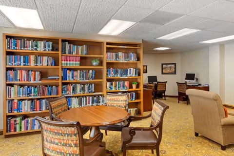 A cozy common room with tall bookshelves, a round wooden table with chairs, armchairs and a couple of computer stations.