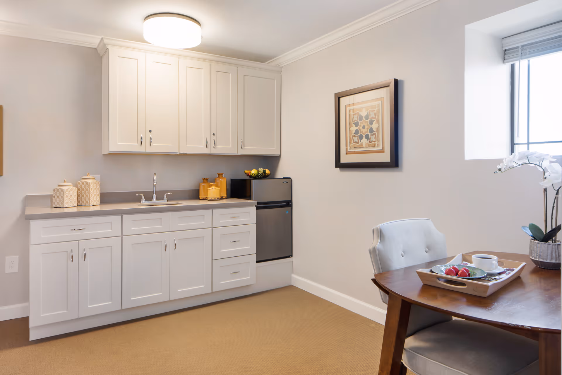 A bright kitchenette with white cabinets, a sink, a mini refrigerator and a small dining table with a chair.