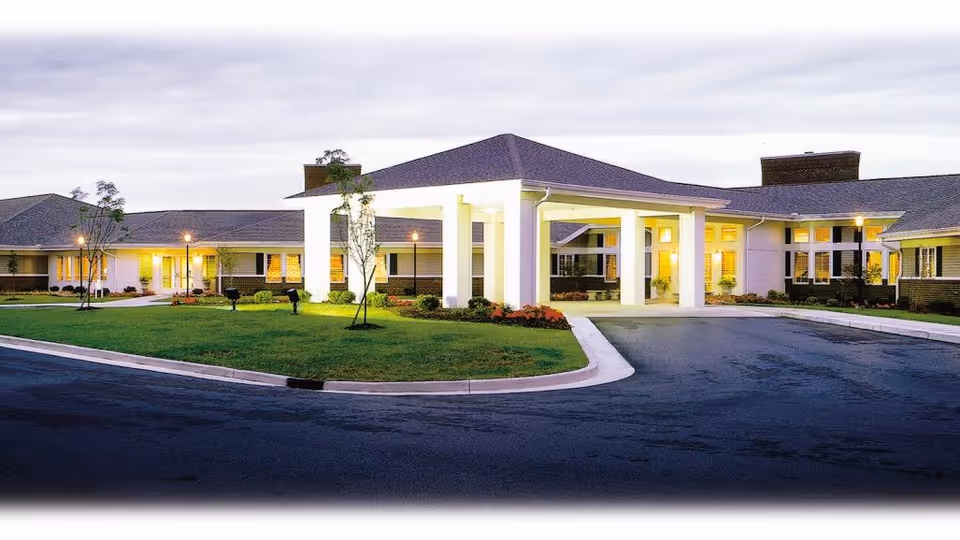 Exterior view of a single-story senior living facility building at dusk with a covered entrance, well-maintained lawn, small trees, and illuminated windows and outdoor lights.
