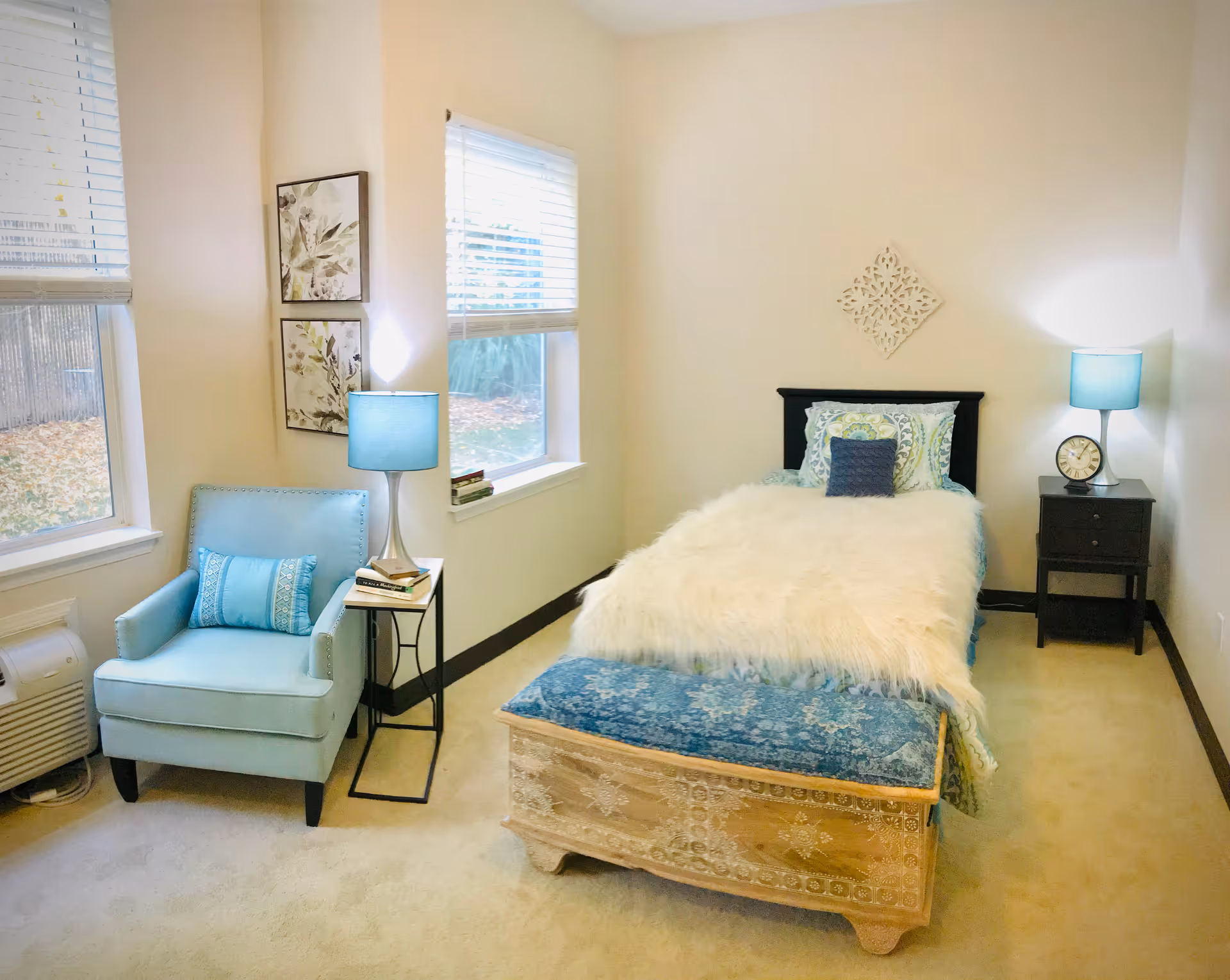 Small furnished bedroom with a twin bed topped with a white faux fur throw, a blue upholstered bench at the foot, teal armchair, side tables and lamps by two windows.