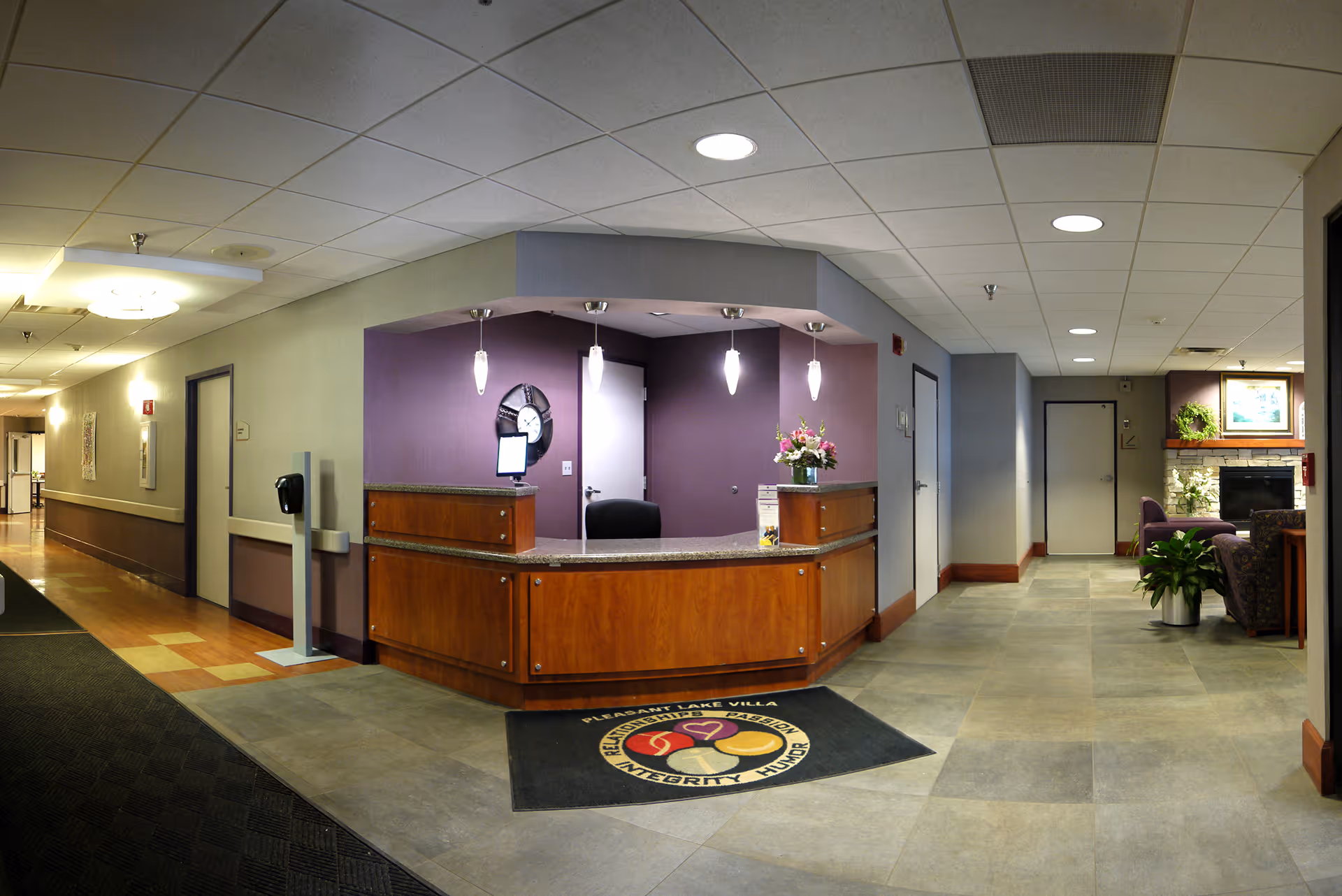Reception area of Pleasant Lake Villa featuring a wooden front desk with a granite countertop, three pendant lights hanging above, a clock on a purple accent wall, and a flower arrangement on the desk. To the right, there is a seating area with armchairs, a fireplace, and framed artwork on the wall. The floor is tiled, and the ceiling has recessed lighting.