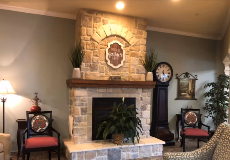 A cozy living room area with a stone fireplace in the center. On the mantel above the fireplace are two potted plants and a decorative sign that reads 'Friends gather here.' To the left and right of the fireplace are chairs with red cushions and decorative backs. A tall grandfather clock and a framed painting hang on the wall to the right. A floor lamp and a small table with a red vase are on the left side. A patterned armchair is partially visible in the foreground on the right.
