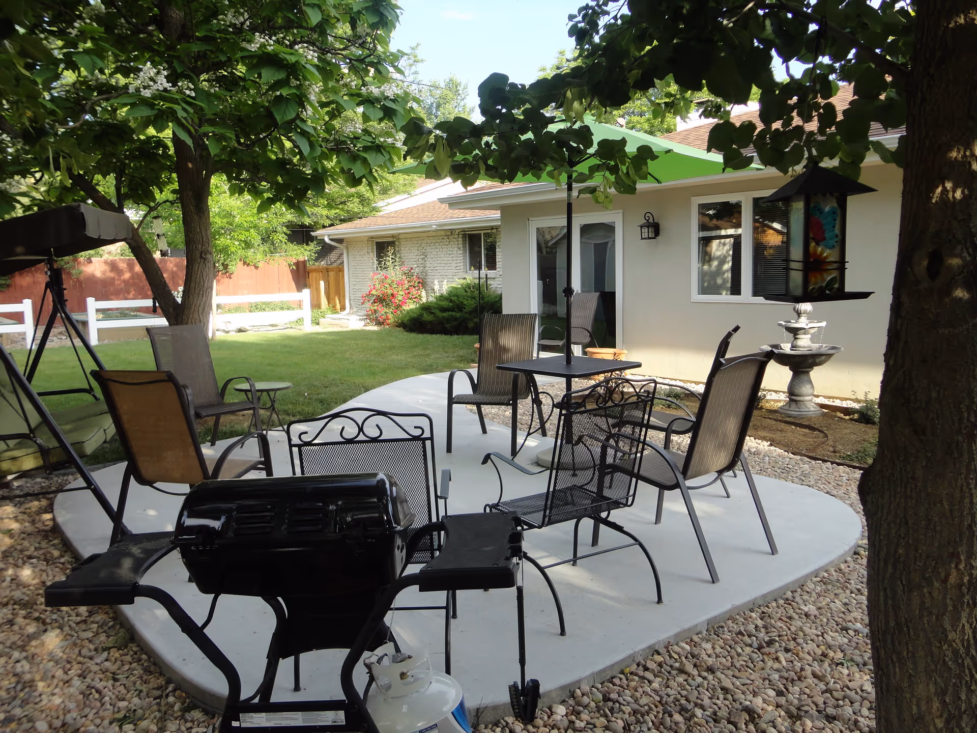Outdoor patio area with a concrete seating space featuring multiple metal and mesh chairs, a table with a green umbrella, a black grill, and a bird feeder hanging from a tree. The area is surrounded by grass, trees, and a white fence, with a building in the background.