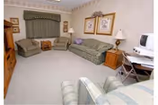 A cozy living room with a green patterned sofa, two matching armchairs, a wooden side table with a lamp, framed artwork on the wall, a window with a valance, and a small desk with a computer and chair.