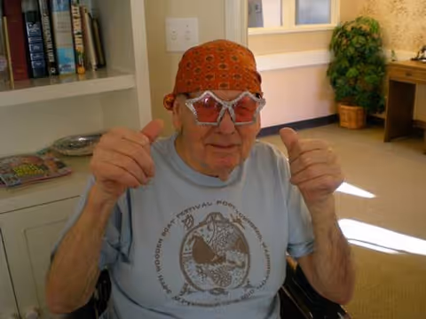 An elderly man wearing a red bandana and star-shaped novelty glasses is sitting indoors, giving two thumbs up. Behind him is a bookshelf with books and magazines, a potted plant, and a desk with a chair in a well-lit room.