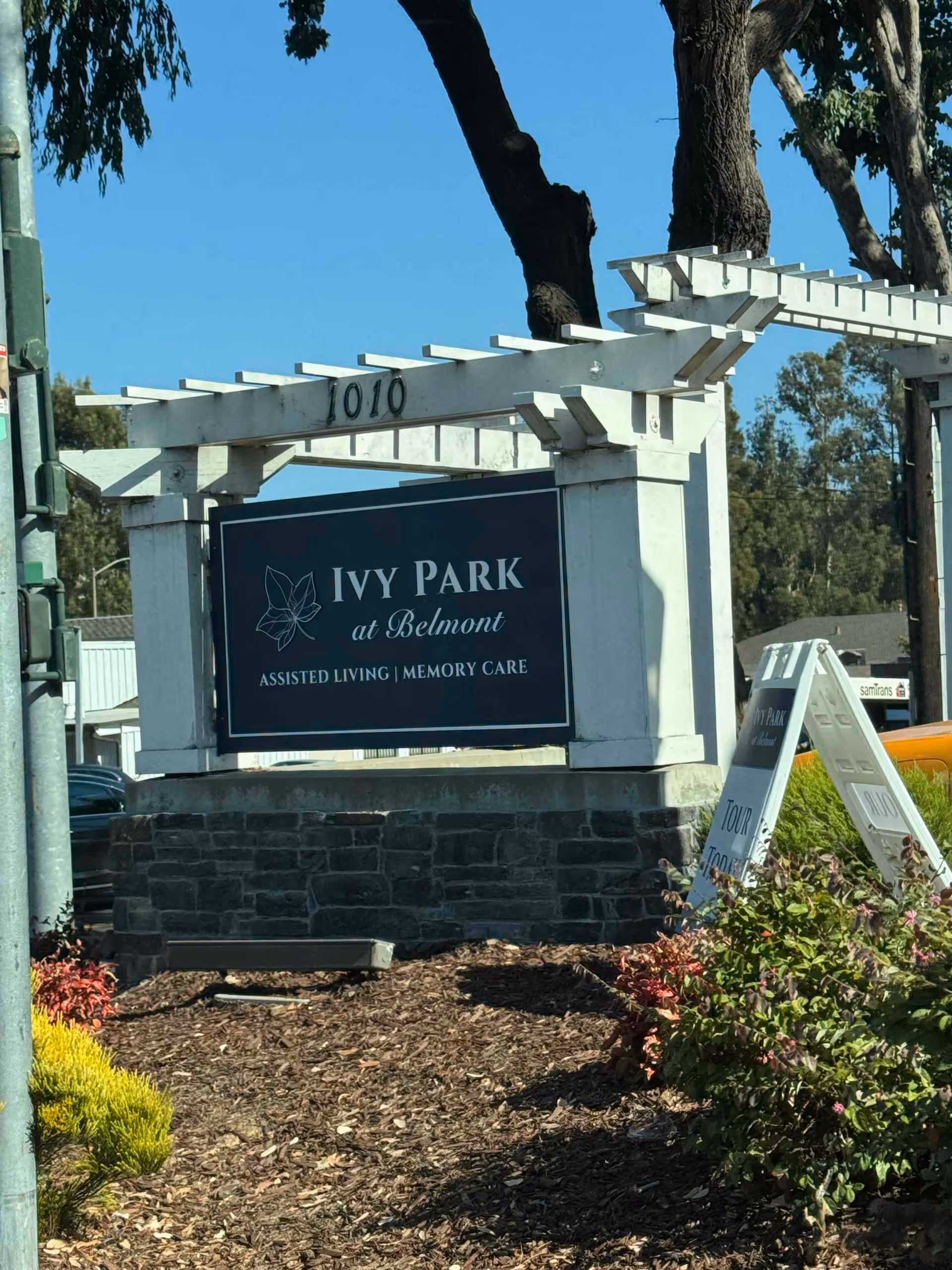 Outdoor sign for Ivy Park at Belmont, an assisted living and memory care facility, mounted on a white wooden pergola structure with the address number 1010 above it. There are trees and bushes around the sign under a clear blue sky.