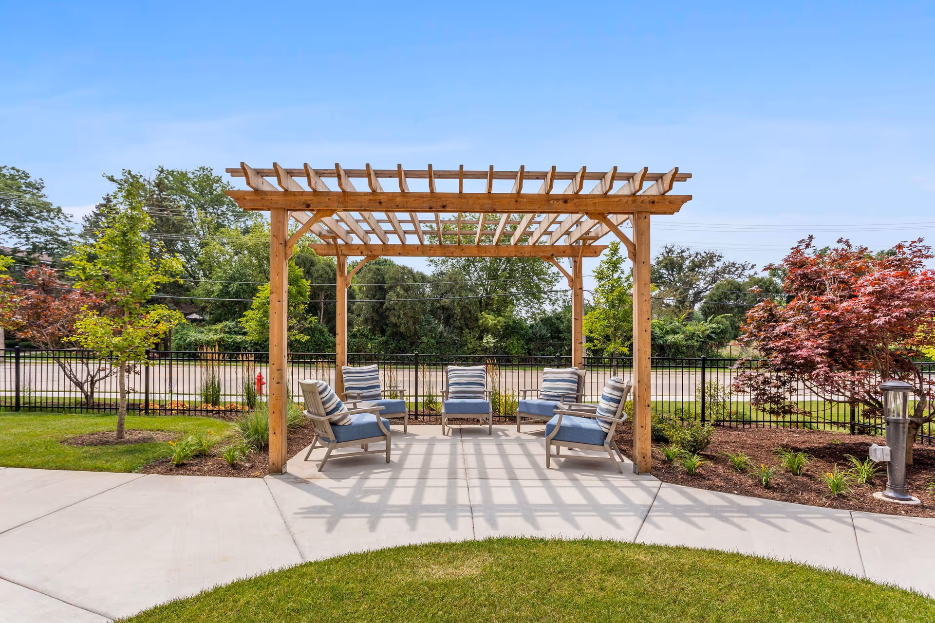 Outdoor seating area with a wooden pergola providing partial shade over five cushioned chairs arranged in a circle on a concrete patio. The area is surrounded by a black metal fence, green grass, and landscaped plants and trees under a clear blue sky.