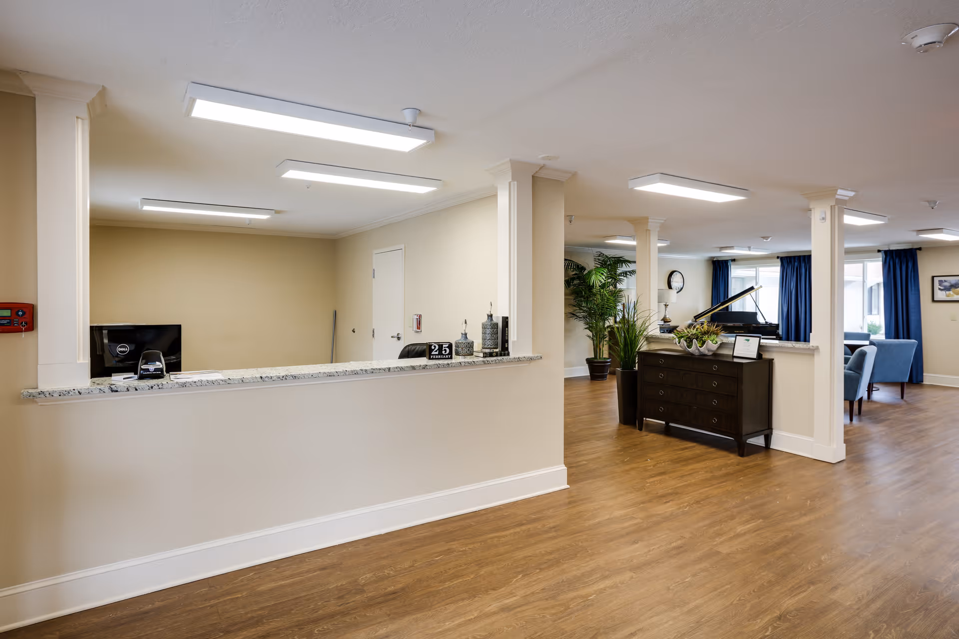 Interior view of a senior living facility reception area with a granite countertop desk, computer, and decorative items. The space has wooden flooring, beige walls, white columns, and ceiling lights. In the background, there is a black piano, plants, a clock on the wall, and blue curtains with seating areas.