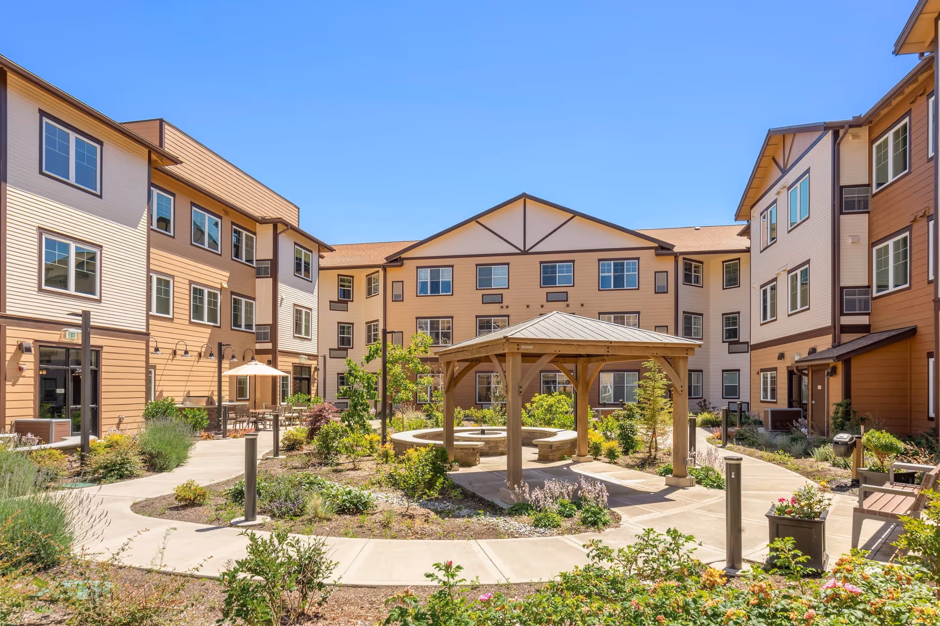 Outdoor courtyard area of Quail Park of Lynnwood featuring a wooden gazebo with a circular stone bench inside, surrounded by landscaped garden beds and paved walkways. The courtyard is enclosed by a three-story building with beige and brown siding under a clear blue sky.