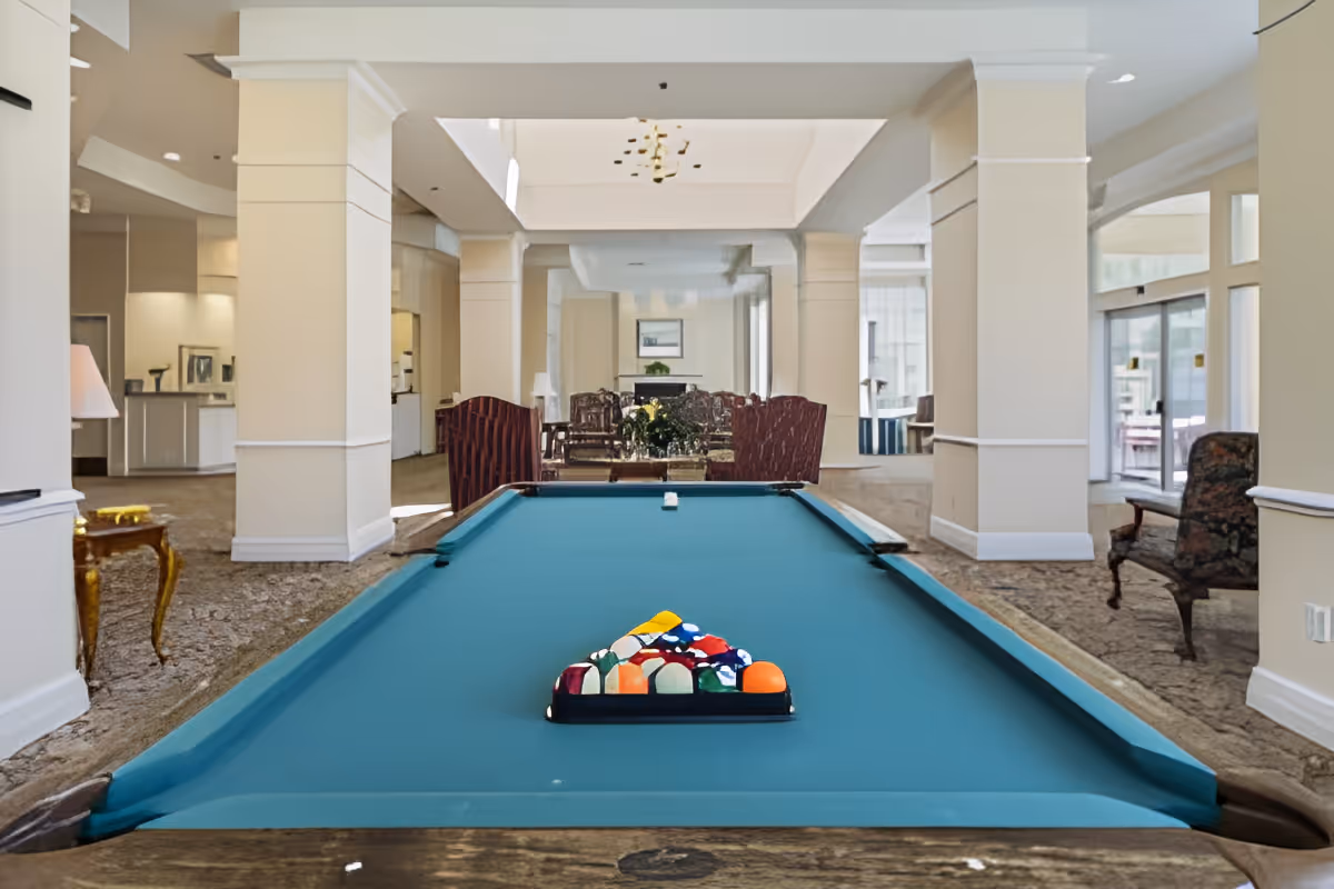 Interior view of a senior living facility lounge area featuring a pool table with a rack of billiard balls set up for a game. The room has beige walls, carpeted floors, and several seating areas with chairs and tables in the background. Large windows allow natural light to enter the space.