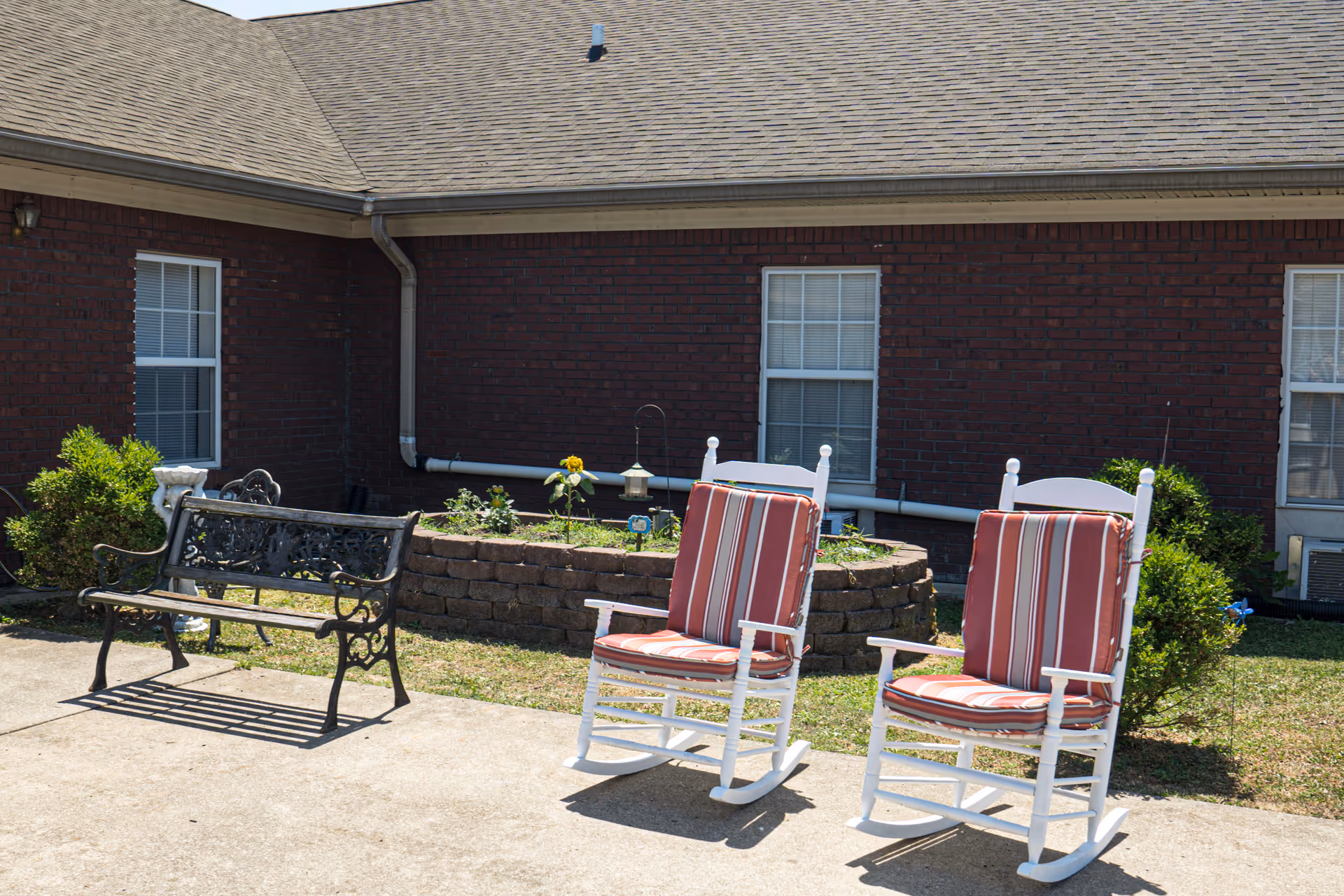 Outdoor patio area with two white rocking chairs with red and white striped cushions and a black metal bench. Behind the seating area is a raised brick flower bed with some plants and a bird feeder, set against a brick building with windows.