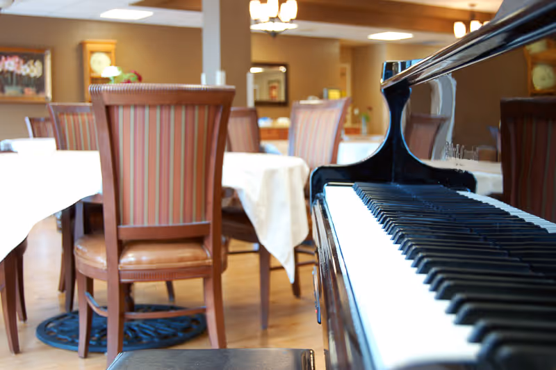 A grand piano keyboard in the foreground with a dining area of tables covered in white cloths and striped-back chairs in the background.