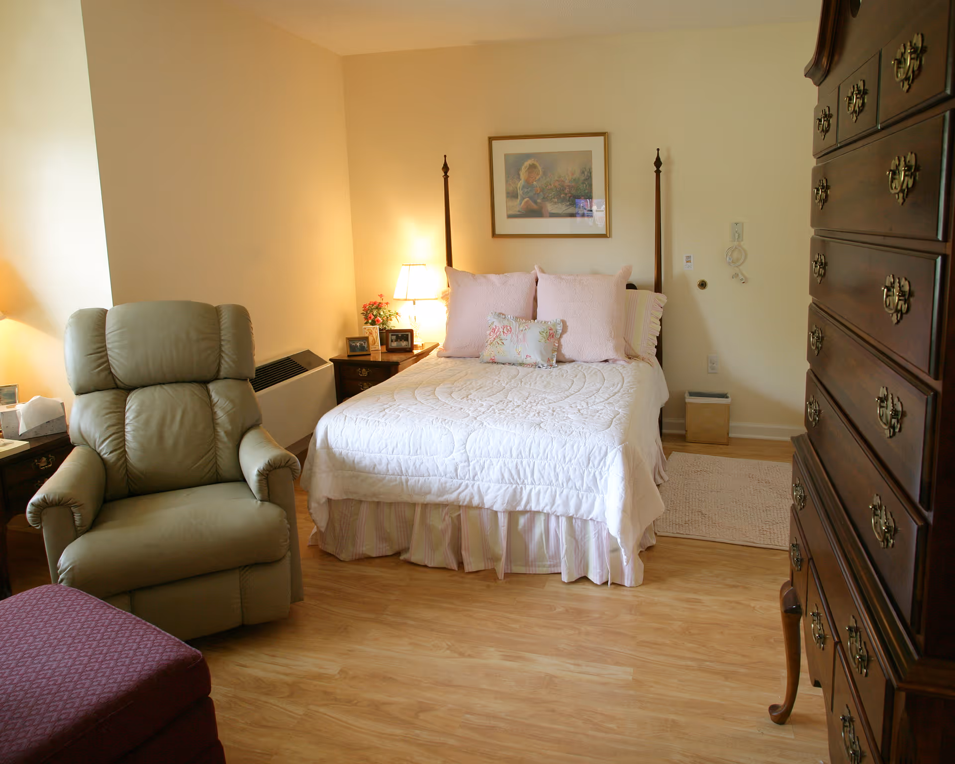 A cozy bedroom in a senior living facility featuring a bed with white and pastel bedding, a green recliner chair, a wooden dresser with brass handles, a nightstand with a lamp and framed photos, and a framed picture on the wall above the bed.
