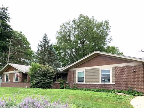 Exterior view of a single-story brick building with brown siding accents, surrounded by green grass, bushes, and tall trees under a cloudy sky.
