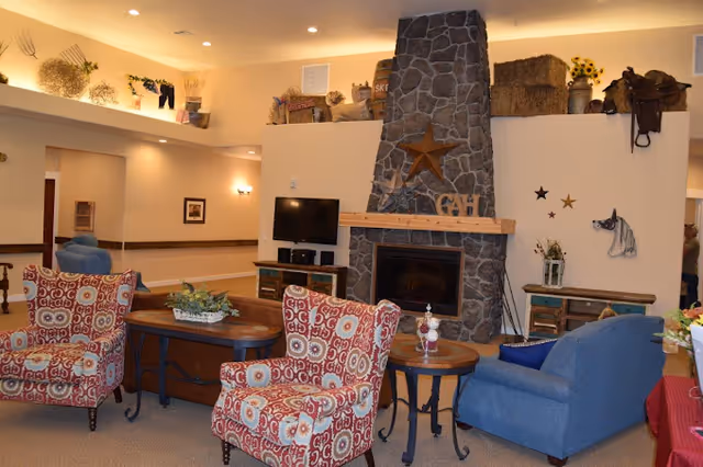 A cozy living room area in a senior living facility featuring a stone fireplace with a wooden mantel decorated with a star and letters. The room has patterned armchairs, a blue armchair, wooden tables, a TV on a stand, and various rustic decorations including baskets, sunflowers, and a horse saddle on a high shelf.