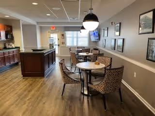 A dining area in a senior living facility with round tables and patterned chairs. The room has wood flooring, neutral-colored walls with framed pictures, a mounted TV, and a counter area with cabinets. Ceiling lights and a large window provide illumination.
